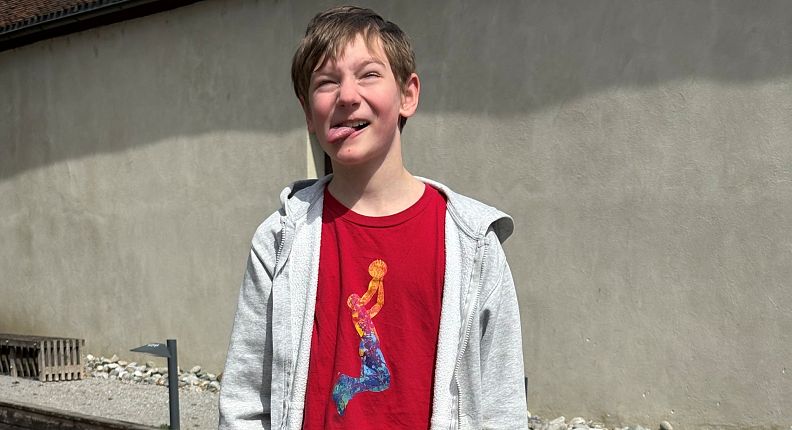 Ein Junge steht draußen vor einer Wand, trägt ein rotes T-Shirt mit einem bunten Motiv und eine graue Kapuzenjacke. Die Sonne scheint, und er blinzelt leicht.