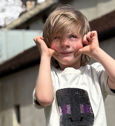Ein Kind mit blonden Haaren steht draußen und macht mit den Händen eine lustige Gesichtsbewegung. Es trägt ein T-Shirt mit einem schwarzen Motiv.