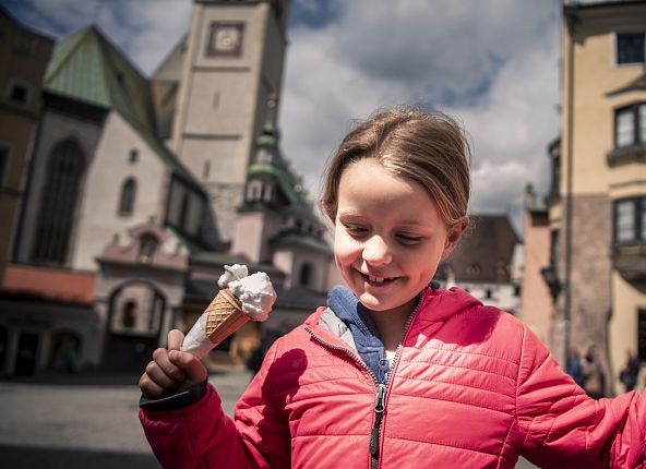 Mädchen in pinker Jacke hält lächelnd ein Eis vor einer historischen Stadtkulisse mit Kirchturm.