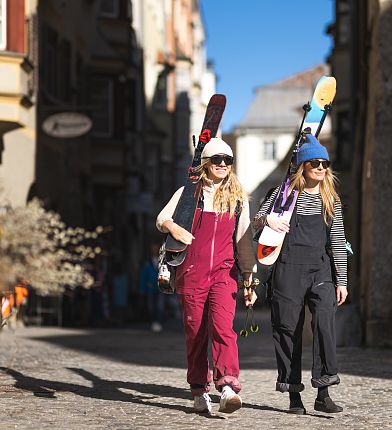 Zwei Personen in Skikleidung tragen Skier auf den Schultern und spazieren bei Sonnenschein durch eine gepflasterte Straße.