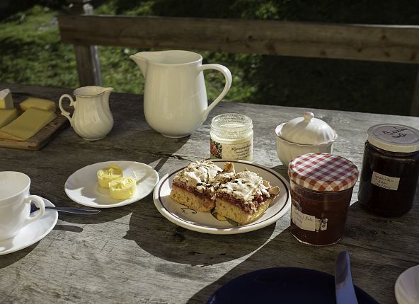 Auf einem Holztisch wird eine Tiroler Almjause mit Milch, Butter, Käse, Marmeladen und Kuchen serviert.