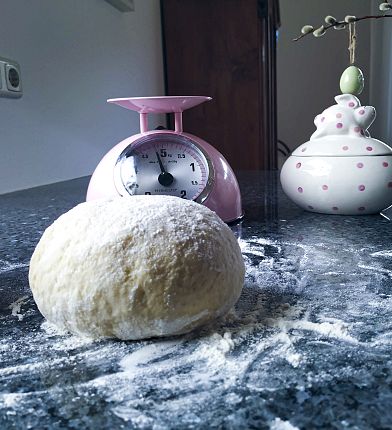 Ein Teigball liegt auf einer bemehlten Oberfläche, mit einer rosa Waage und einem dekorativen Gefäß im Hintergrund.