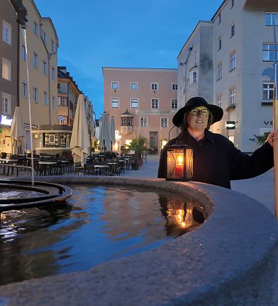 Eine Person mit Laterne und Stab in altem Gewand und Hut steht vor einem an der Wand hängenden Brunnen. Sie symbolisiert die Nachtwächter, welche in mittelalterlichen Städten allgegenwärtig waren.