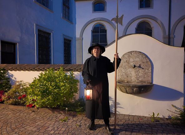 Eine Person mit Laterne und Stab in altem Gewand und Hut steht vor einem an der Wand hängenden Brunnen. Sie symbolisiert die Nachtwächter, welche in mittelalterlichen Städten allgegenwärtig waren.
