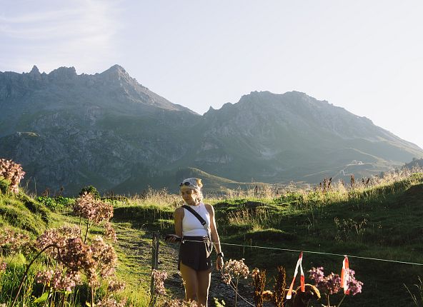 Eine Frau wandert in einer sonnigen Berglandschaft. Im Hintergrund erheben sich hohe Berggipfel unter einem klaren Himmel.