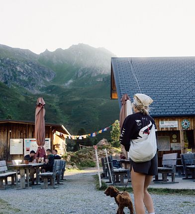 Eine Berghütte vor Bergkulisse, Menschen sitzen an Holztischen. Eine Person mit Rucksack im Vordergrund. Berglandschaft und Sonnenschirme sichtbar.