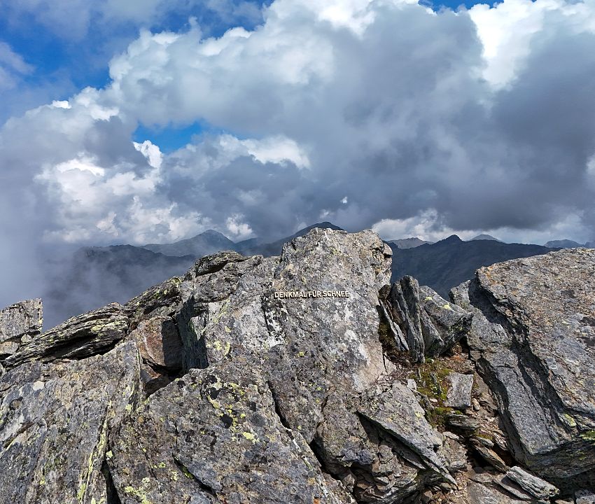 Bergspitzen mit dem Messingschriftzug Denkmal für Schnee, darüber Wolken und etwas blauer Himmel