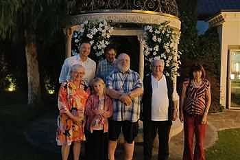 Eine Gruppe von sieben Menschen, unterschiedlichen Alters, posiert draußen vor einem dekorierten Pavillon mit Blumen, abends oder bei Dämmerung. Einige lächeln in die Kamera.