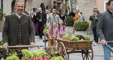 Zwei Männer in traditioneller Kleidung ziehen Holzkarren mit Gemüse durch eine enge Altstadtgasse, flankiert von schaulustigen Passanten.