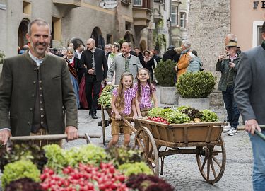 Zwei Männer in traditioneller Kleidung ziehen Holzkarren mit Gemüse durch eine enge Altstadtgasse, flankiert von schaulustigen Passanten.