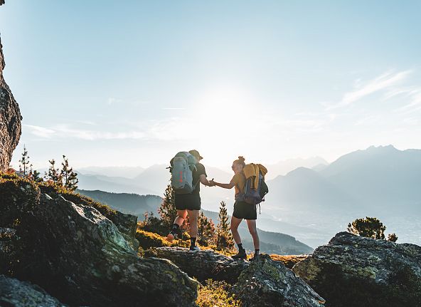 Zwei Menschen wandern auf einem Bergpfad bei Sonnenaufgang, umgeben von Bergen und Bäumen. Einer hilft dem anderen über einen Felsen.