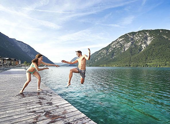 Zwei Personen auf einem Steg an einem Bergsee: Ein Mann springt ins Wasser, während eine Frau ihn lachend ansieht. Die Berge spiegeln sich im klaren Wasser.