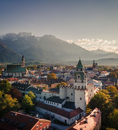 Eine idyllische Stadt mit historischen Gebäuden und Kirchtürmen, umgeben von grünen Bäumen und Bergen, bei Sonnenuntergang im Hintergrund.