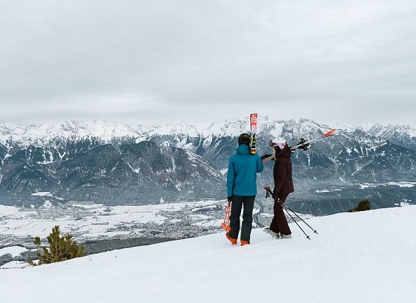 Zwei Skifahrer betrachten von einer Berghöhe aus eine schneebedeckte Alpenlandschaft. Wolkenverhangener Himmel verstärkt die winterliche Atmosphäre.