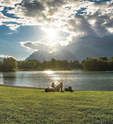 Zwei Personen sitzen auf einer Wiese am Seeufer, umgeben von Natur, während die Sonne hinter Wolken hervorscheint.