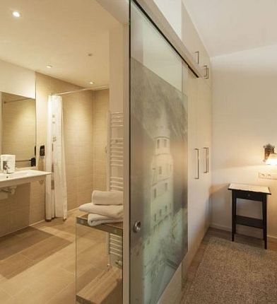 Hotelzimmer mit hellem Bett, Nachttischlampe und Glaswand trennt Badezimmer mit Dusche und Waschbecken ab.
