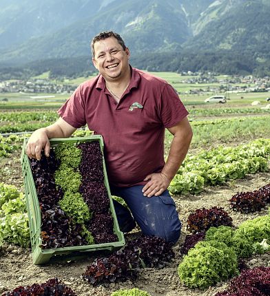 Ein Bauer kniet auf einem Salatfeld, lächelt und hält eine Kiste mit frischem Salat. Im Hintergrund sind eine Landschaft und Berge sichtbar.