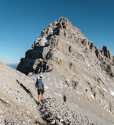 Ein Wanderer auf einem steinigen Bergpfad mit einem großen Berg im Hintergrund unter einem klaren blauen Himmel.