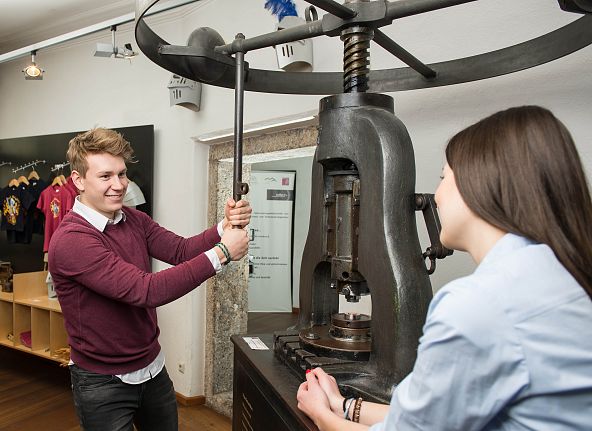 Junger Mann an einer alten Druckerpresse, beobachtet von einer Frau in einem Museumsraum.