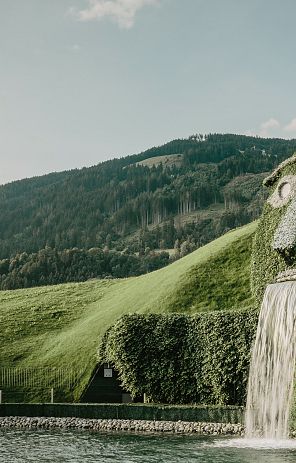 Pflanzenbedecktes Gesicht eines Riesen mit Wasser, das aus dem Mund fließt, vor einer Berglandschaft mit grünen Hügeln.