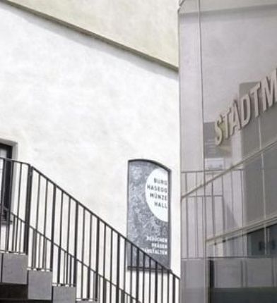 Eingang eines Stadtmuseums mit Treppen, Glasfassade und Schildern, die auf das Museum hinweisen, neben historischen Mauern.