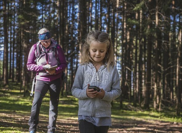 Ein Kind und ein Erwachsener navigieren mit Geräten durch einen sonnigen Wald. Beide tragen Freizeitkleidung und genießen die Natur.