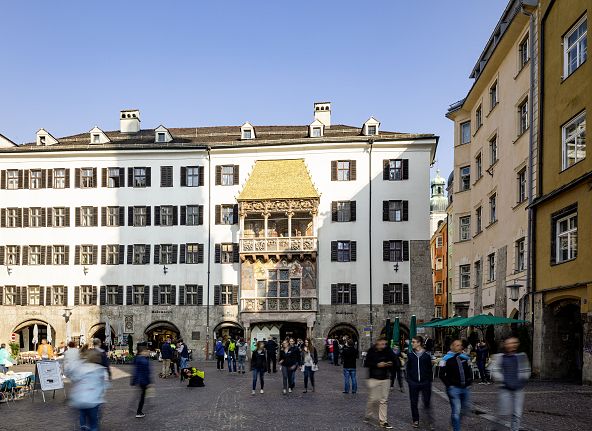 Menschen flanieren durch Innsbrucks Altstadt unter dem Goldenen Dachl. Historische Gebäude umgeben den sonnigen Platz mit zahlreichen Besuchern.