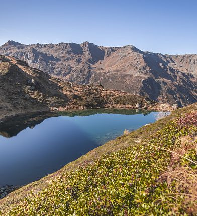 Ein klarer Bergsee in den Alpen, umgeben von Bergen und herbstlicher Vegetation, die sich in den Wasseroberfläche spiegelt.