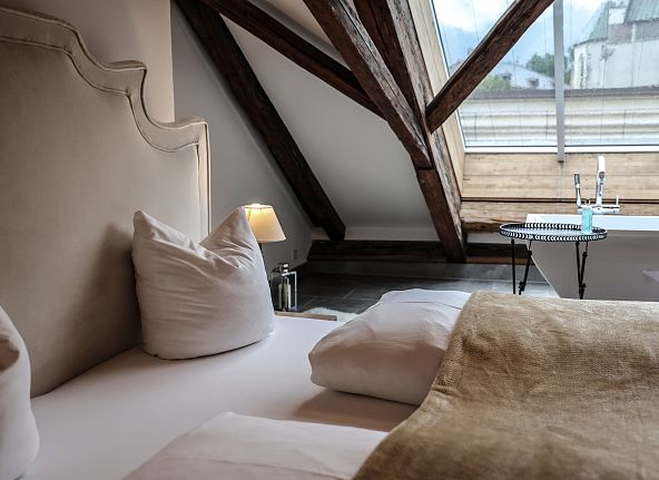 Gemütliches Schlafzimmer unter dem Dach mit sichtbaren Holzbalken, einem großen Bett und einem Dachfenster, das viel Licht einlässt.