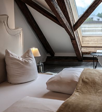 Schlafzimmer mit Dachschräge, Holzbalken, großem Fenster, Bett mit Kissen und Decke.