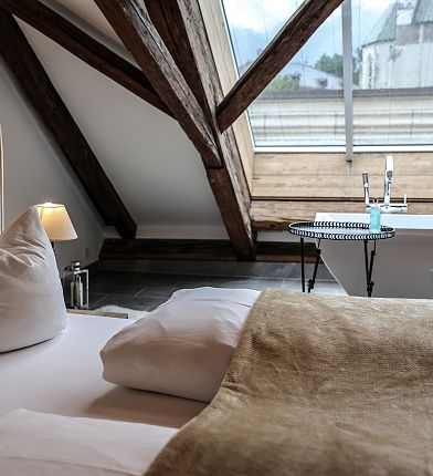Schlafzimmer im Dachgeschoss mit schräge Holzdecke, weichem Bett, Tisch mit Lampe und großem Fenster mit Ausblick auf die Berge.