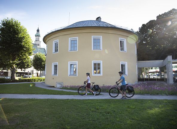 Zwei Radfahrer passieren ein rundes, gelbes Gebäude in einer grünen Landschaft. Im Hintergrund ist ein Kirchturm zu sehen, umgeben von Bäumen.