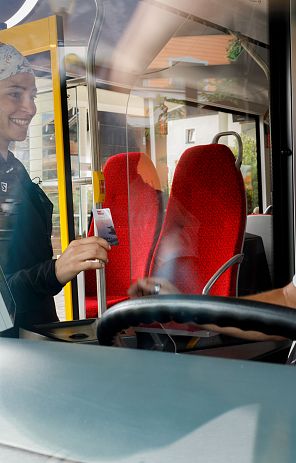 Frau zeigt lächelnd ihr Ticket einem Busfahrer durch eine Trennscheibe. Beide sind in einem modernen Bus, die Stimmung wirkt freundlich.