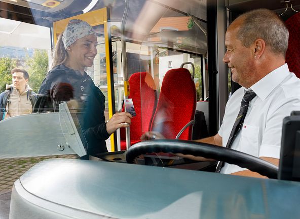 Frau zeigt lächelnd ihr Ticket einem Busfahrer durch eine Trennscheibe. Beide sind in einem modernen Bus, die Stimmung wirkt freundlich.