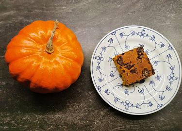 Ein Kürbis und ein Stück Kürbiskuchen auf einem Teller mit blauem Muster, auf dunklem Steinuntergrund.