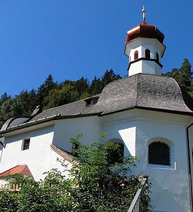 Weiße Kapelle mit rotem Turmdach vor blauem Himmel, umgeben von Bäumen.