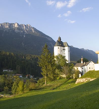 Ein malerisches Schloss mit einem hohen Turm und einem majestätischen Berg im Hintergrund. Die Szene ist von grünen Hügeln und bewaldeter Landschaft umgeben.