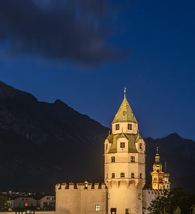 Ein beleuchteter Turm in Innsbruck bei Nacht mit Bergen im Hintergrund vor einem tiefblauen Himmel.
