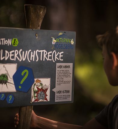 Ein Junge steht vor einem Schild mit der Aufschrift "Bildersuchstrecke", das mit Bildern und Fragen versehen ist.