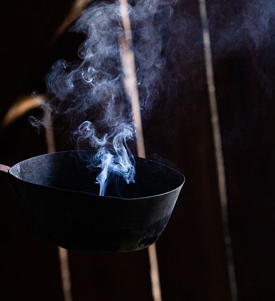 Schwarze Räucherschale mit aufsteigendem Rauch vor dunklem, holzgetäfeltem Hintergrund.