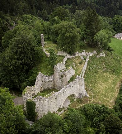 Eine Burgruine in einem grünen Waldgebiet, nahe einer kleinen Kirche mit spitzem Turm, umringt von hohen Bäumen.