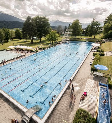 Großes Schwimmbad im Freien mit Schwimmern. Umgeben von Bäumen, Wiesen und Bergen im Hintergrund. Der Himmel ist leicht bewölkt.