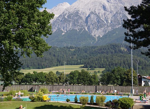 Ein Freibad mit Menschen im Vordergrund und eine schöne Berglandschaft im Hintergrund. Der Pool ist von Bäumen und einer Wiese umgeben.