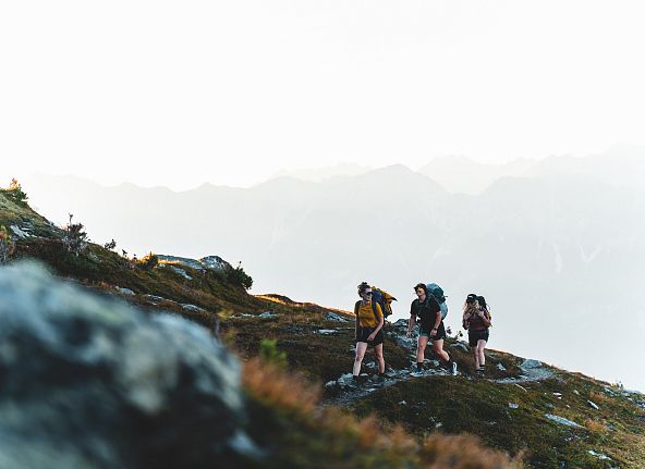 Drei Wanderer auf einem Bergpfad mit Rucksäcken und Trekkingkleidung, vor einer beeindruckenden Bergkulisse und bewölktem Himmel.