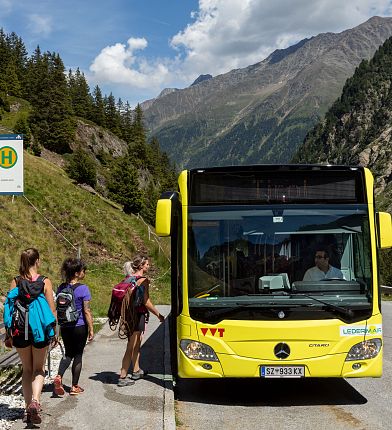 Ein gelber Bus steht an einer Haltestelle in den Bergen. Drei Menschen warten, um einzusteigen. Im Hintergrund sind grüne Bäume und hohe Berge erkennbar.