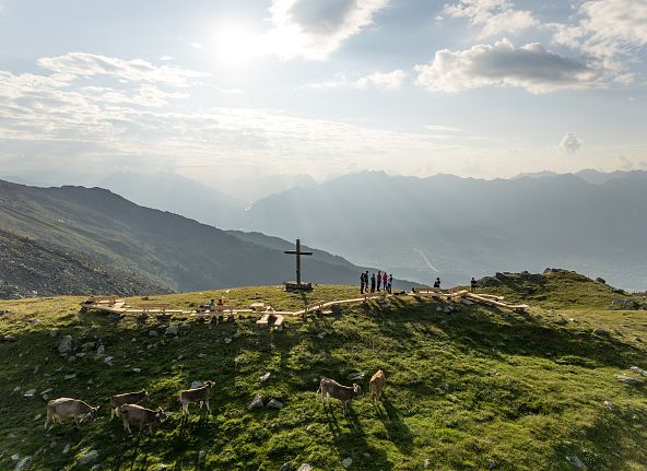Alpine Landschaft mit Ziegen auf einer Wiese, einem Holzkreuz und Bergen im Hintergrund. Der Himmel ist teilweise bewölkt.