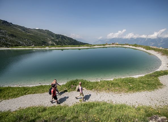 Zwei Wanderer spazieren an einem klaren Bergsee entlang, umgeben von grüner Natur und einem blauen Himmel mit Bergblick im Hintergrund.