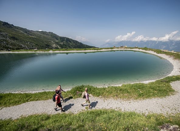 Familie beim Wandern an einem Bergsee mit grünen Hügeln und blauem Himmel im Hintergrund.