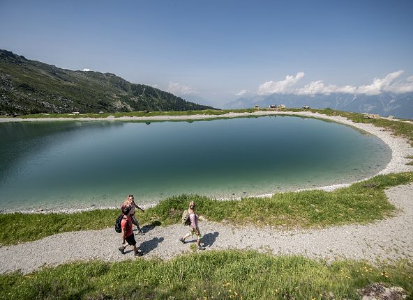 Drei Wanderer spazieren an einem runden Bergsee, der von Wiesen und Berglandschaft umgeben ist. Im Hintergrund erstrecken sich Berggipfel und ein blauer Himmel.
